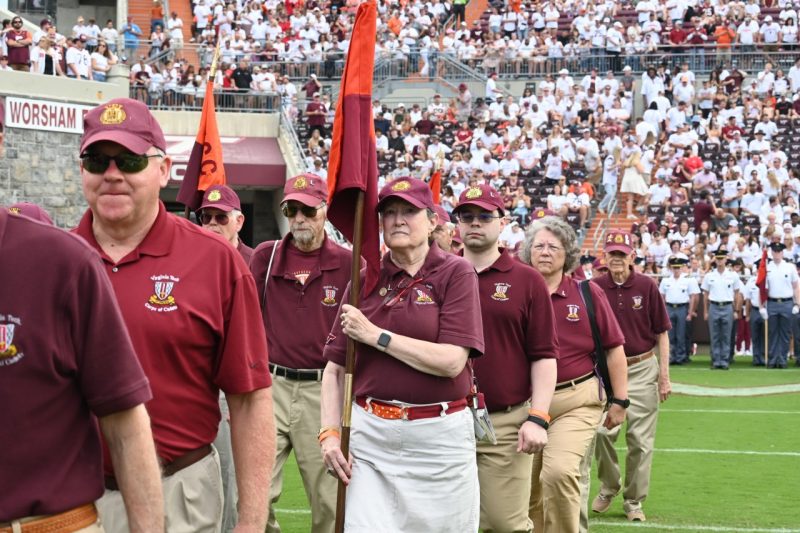 Men and women alumni march onto a football field carrying guidons. They are wearing matching maroon polo shirts bearing a Corps of Cadets logo.